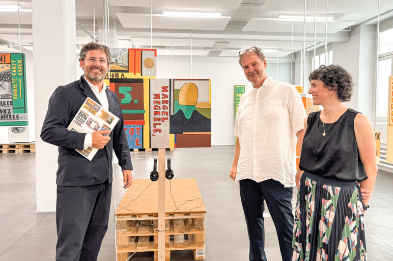 Dr. Marcel Megerle, Till Velten und Patrizia Keller stehen in einem gro&szlig;en Raum. Im Hintergrund sind bunte Kunstwerke zu sehen.
