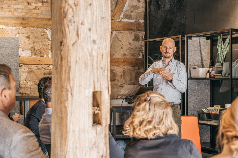 Mehrere Menschen sitzen in einem Raum mit dem R&uuml;cken zur Kamera und schauen zu Sebastian Fittko, der rechts im Bild steht und einen Vortrag h&auml;lt
