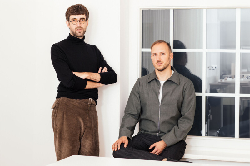 Steffen Vogt lehnt an einer wei&szlig;en Wand, neben ihm sitzt Romas Stukenberg auf einem Fensterbrett, beide schauen direkt in die Kamera