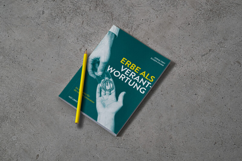 Das Buch "Erbe als Verantwortung" liegt auf einem Betonboden, darauf ein gelber Bleistift. Auf dem Buchcover sieht man eine &auml;ltere Hand, die einen Kompass in eine j&uuml;ngere Hand legt.