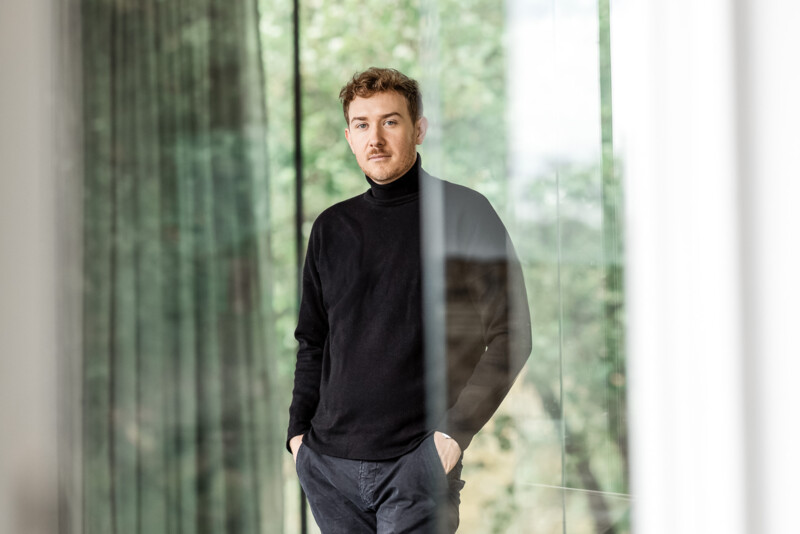 Tristan Horx steht in einem Raum vor einer großen Glasscheibe, vor dem Fenster sind unscharf Bäume zu erkennen. Er hat die Hände in den Hosentaschen und schaut direkt in die Kamera.