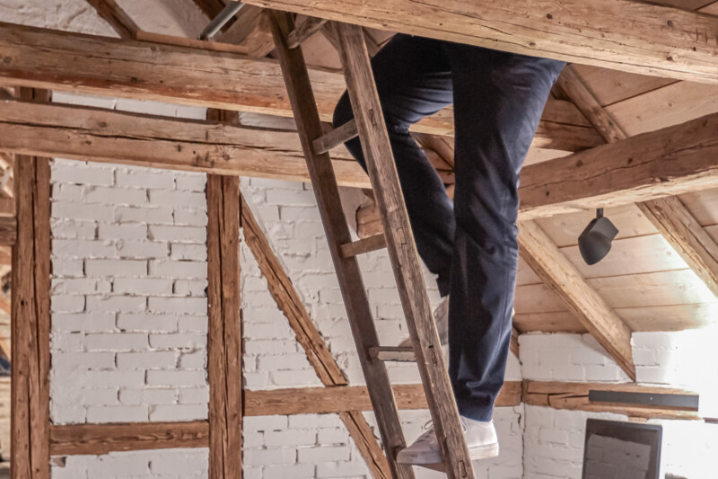 Eine Person steigt eine Holzleiter in einem alten Fachwerkhaus nach oben, man sieht nur noch ihre Beine, der Oberkörper ist außerhalb des Bildes