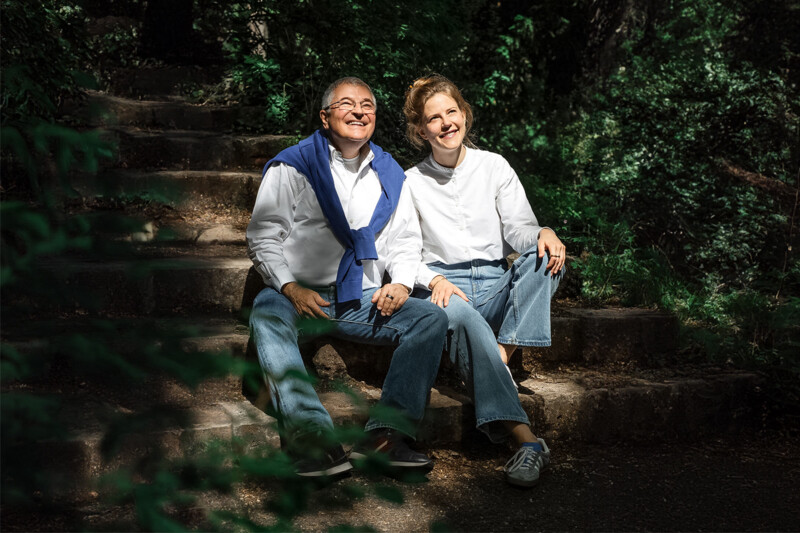 Till Novotny sitzt neben seiner Tochter Leonie Novotny auf Steinstufen, umgeben von Bäumen und Sträuchern. Sie lächeln und blicken nach oben in Richtung Sonne.